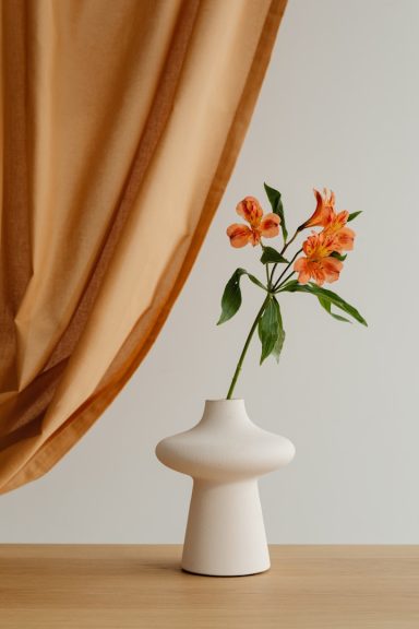 Vase mit orangefarbenen Blumen vor einem braunen Vorhang.